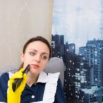 Przemyślana gospodyni w wieżowcu siedząca przed oknem z widokiem na dachy miasta z telefonem komórkowym w dłoni