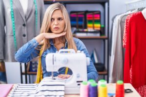 Blondynka projektantka krawcowa używająca maszyny do szycia podcinającej gardło ręką jako nożem grozi agresją z wściekłą przemocą