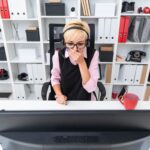 Młoda dziewczyna w słuchawkach siedzi przy stole komputera i zakryła usta dłonią.