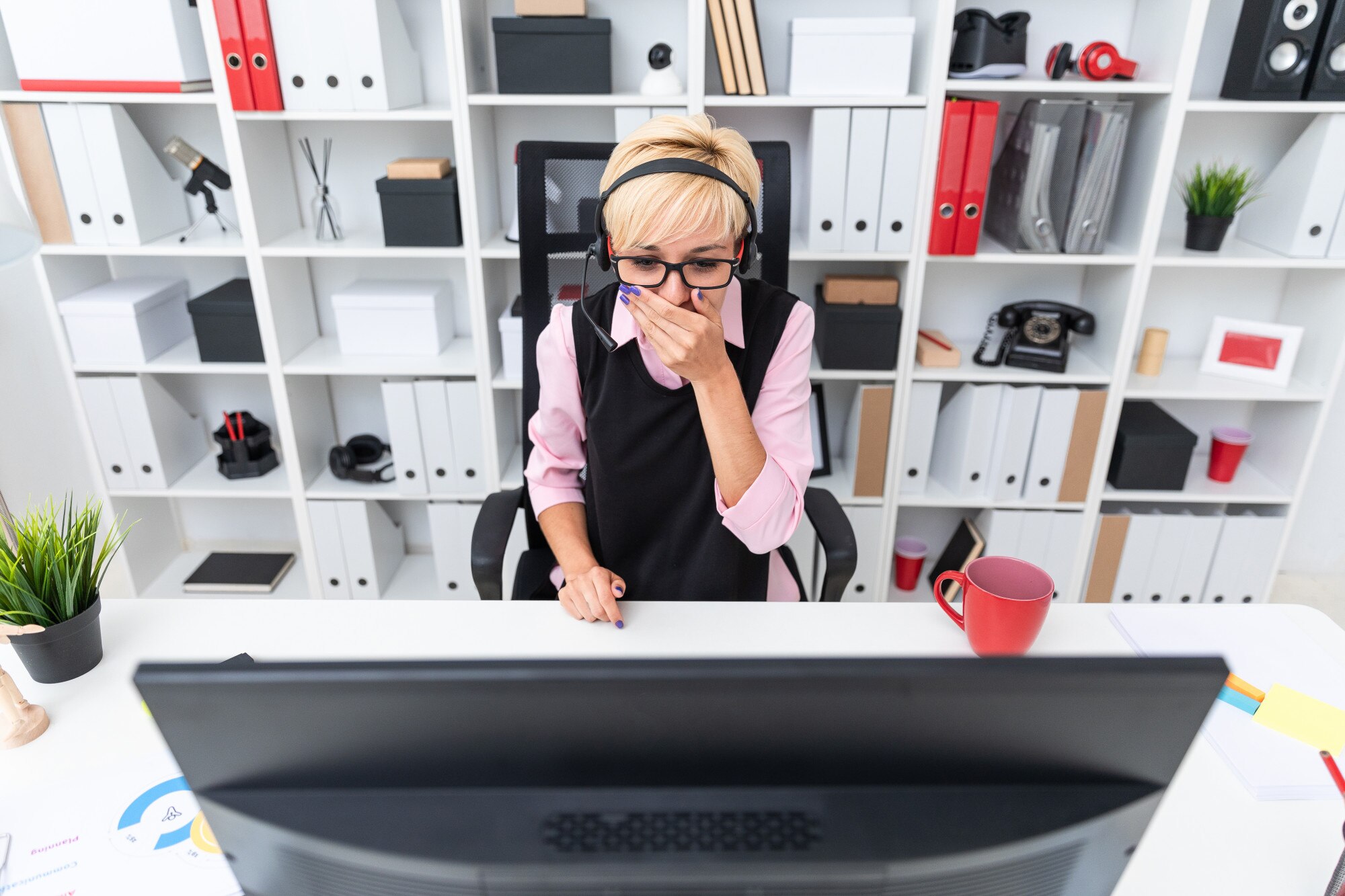 Młoda dziewczyna w słuchawkach siedzi przy stole komputera i zakryła usta dłonią.